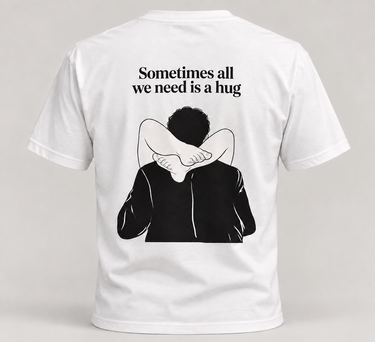 Back of a white tee with Sometimes all we need is a hug in smaller black serif lettering above a cheeky illustration
